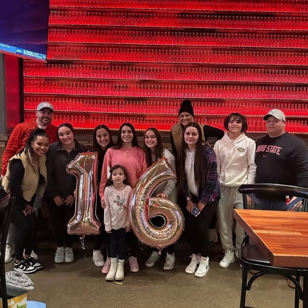 A large group of people, including children, pose with "16" balloons in front of a red-lit wall.