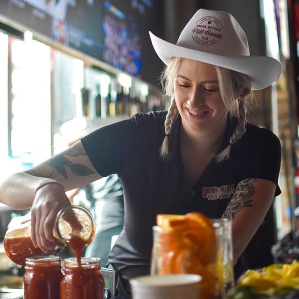 A smiling bartender in a cowboy hat pours a red drink into mason jars at Urban Meyer's Pint House.