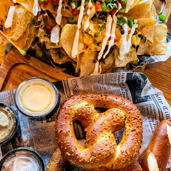 Loaded nachos with sour cream and pretzels with dipping sauces on newspaper-lined trays.