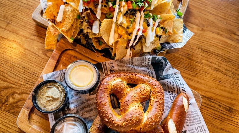 Loaded nachos with sour cream and pretzels with dipping sauces on newspaper-lined trays.