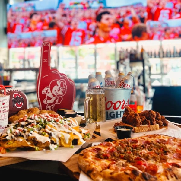 Nachos, pizza, fried chicken, beer, and a Coors Light bucket with a football game on a large screen.