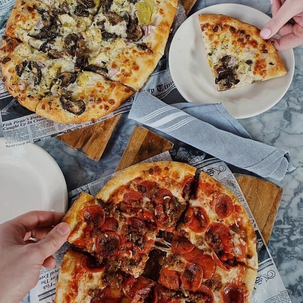 Two hands reaching for slices of a pepperoni pizza and a white pizza with mushrooms and artichokes.