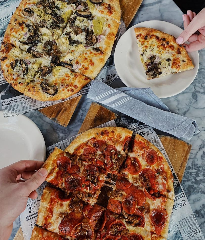 Two hands reaching for slices of a pepperoni pizza and a white pizza with mushrooms and artichokes.