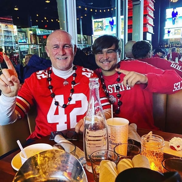 Two smiling men in red Ohio State shirts and beaded necklaces sit at a restaurant table.