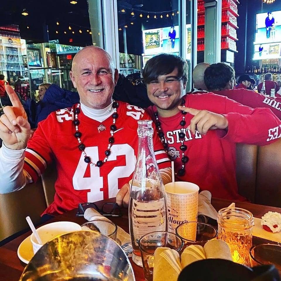 Two smiling men in red Ohio State shirts and beaded necklaces sit at a restaurant table.