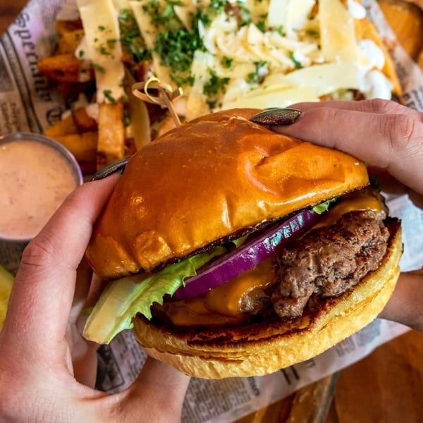 Hands holding a cheeseburger with lettuce and red onion, with fries and a pickle in the background.