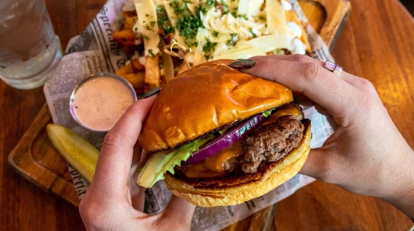 Hands holding a cheeseburger with lettuce and red onion, with fries and a pickle in the background.