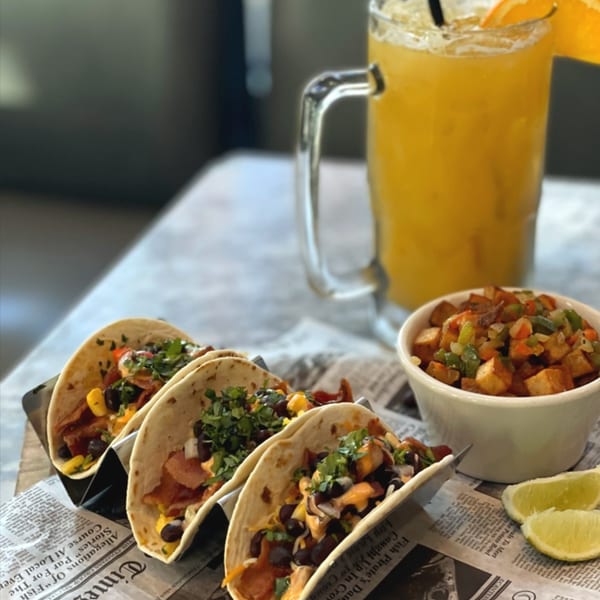 Three tacos, a side of potatoes, and an orange drink on a newspaper-lined tray.