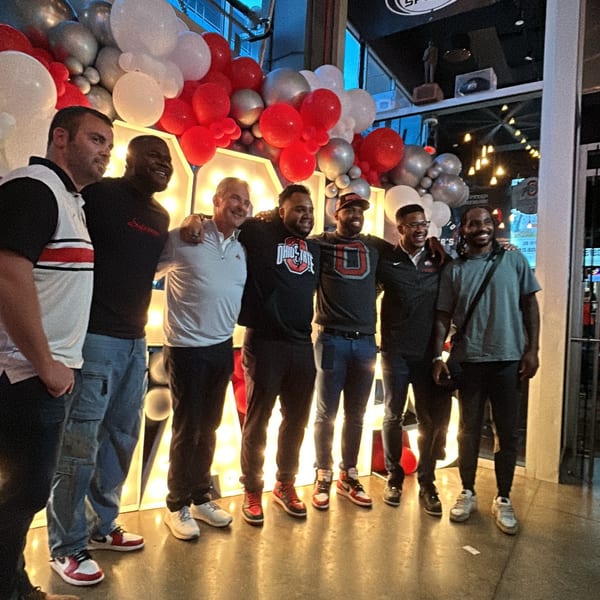 Six men pose smiling in front of a large illuminated sign and red, white, and silver balloons.