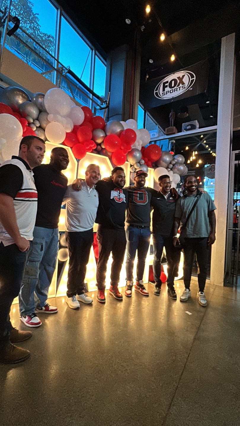Six men pose smiling in front of a large illuminated sign and red, white, and silver balloons.