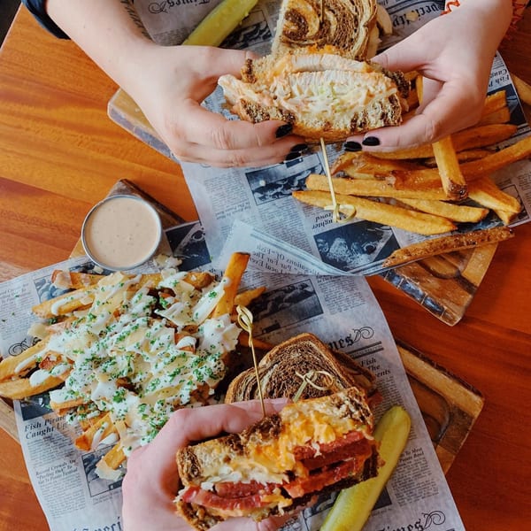 Two people holding sandwiches and fries on newspaper-lined trays, with a side of pickles and sauce.