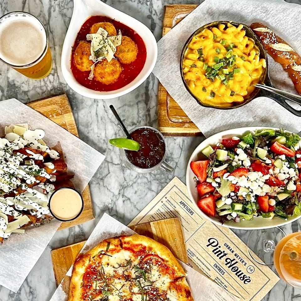 Overhead view of various dishes including pizza, mac and cheese, salad, fries, and drinks on a marble table.