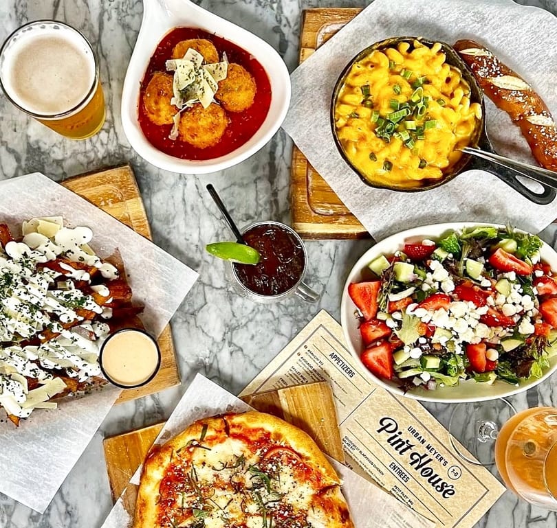 Overhead view of various dishes including pizza, mac and cheese, salad, fries, and drinks on a marble table.