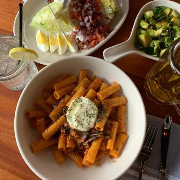 Rigatoni pasta with sauce and a dollop of cream, alongside a wedge salad, brussels sprouts, and drinks.
