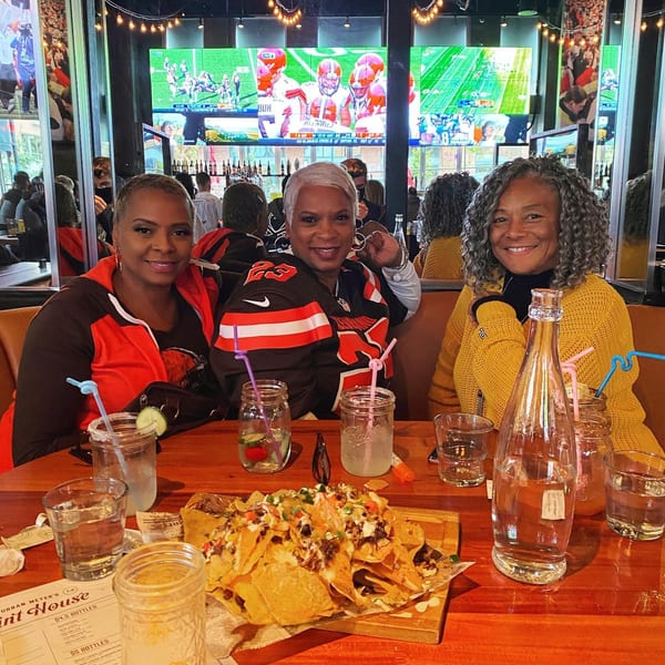 Three women smiling at a table with nachos and drinks, watching football on large screens.