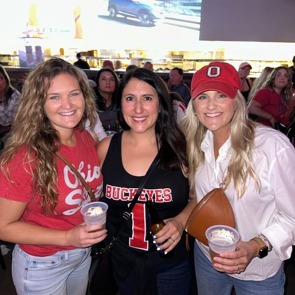 Three smiling women in Ohio State Buckeyes gear holding drinks in a lively bar setting.