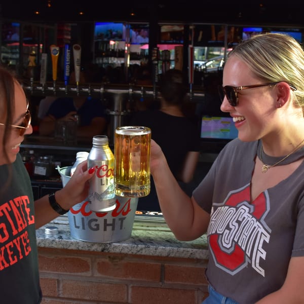 Two women in Ohio State shirts toast with a beer glass and a Coors Light can at a bar.