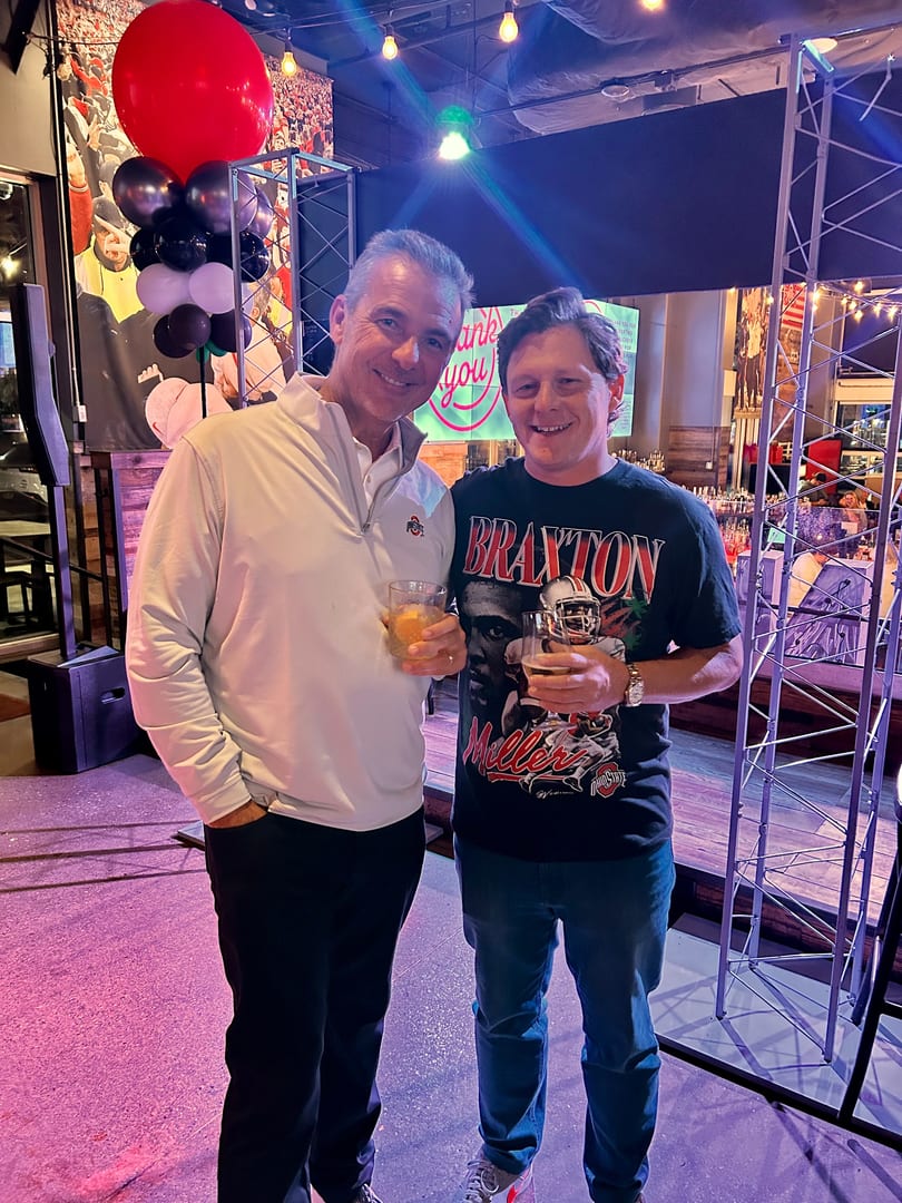 Two men smiling and holding drinks, one wearing an Ohio State shirt, at a lively event.