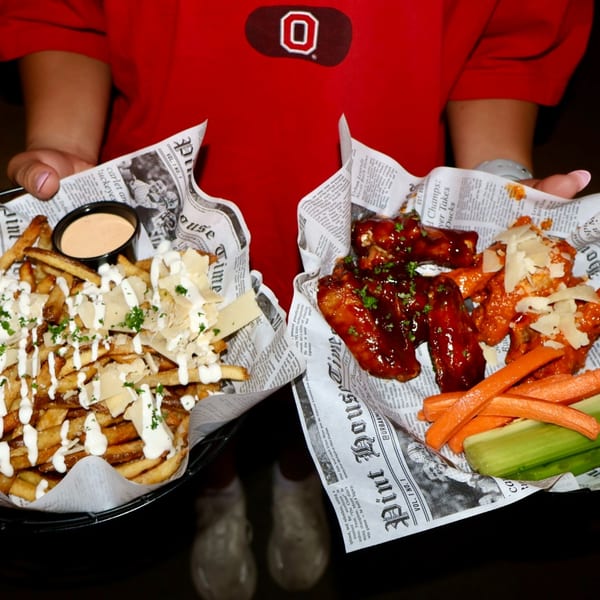 Hands holding two baskets of food: parmesan fries with dipping sauce and chicken wings with vegetables.