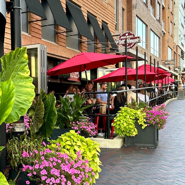 Patrons enjoying outdoor dining under red umbrellas at Urban Meyer's Pint House, surrounded by vibrant flowers.