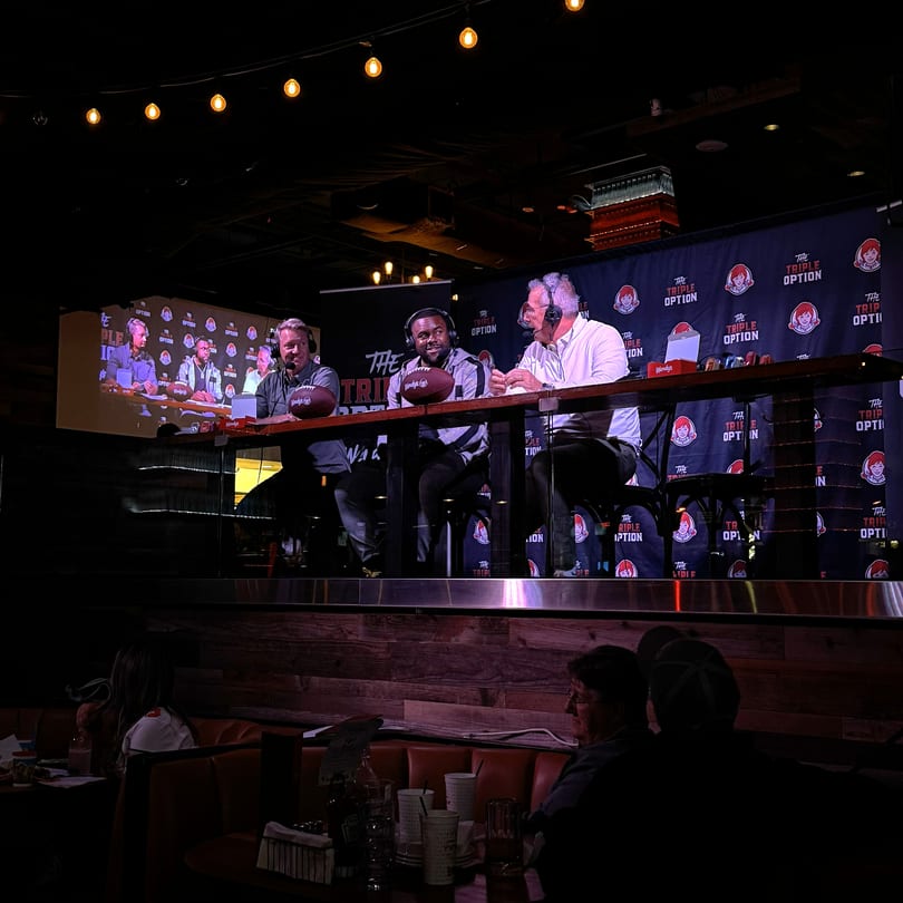 Three men in headsets broadcast "The Triple Option" from a restaurant stage with a Wendy's backdrop.