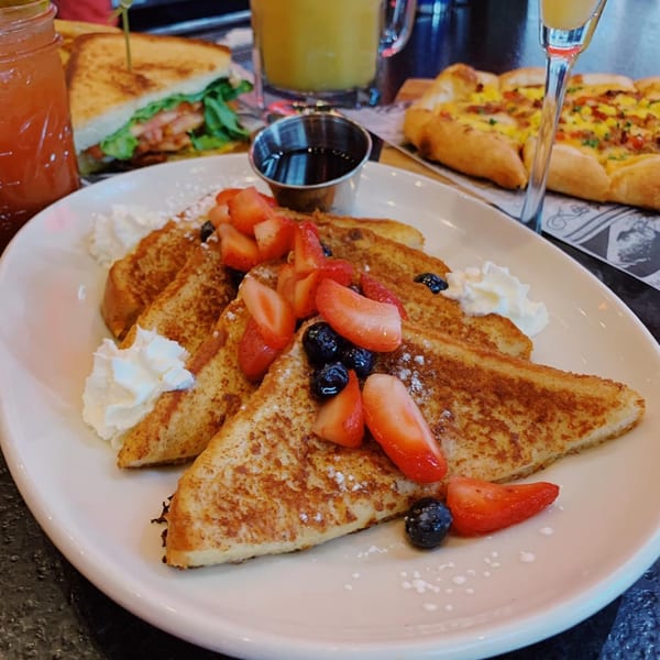 French toast with strawberries, blueberries, whipped cream, and syrup, alongside a sandwich and flatbread.