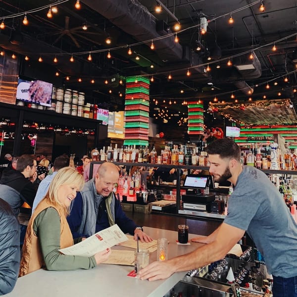 A bartender serves drinks to patrons seated at a lively bar with festive red and green lighting.