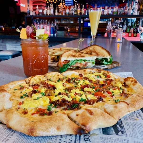 Breakfast pizza, sandwich, fries, Bloody Mary, and mimosa on a bar with bottles in the background.