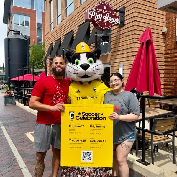 Two people and a cat mascot hold a "Soccer Celebration" sign outside Urban Meyer's Pint House.