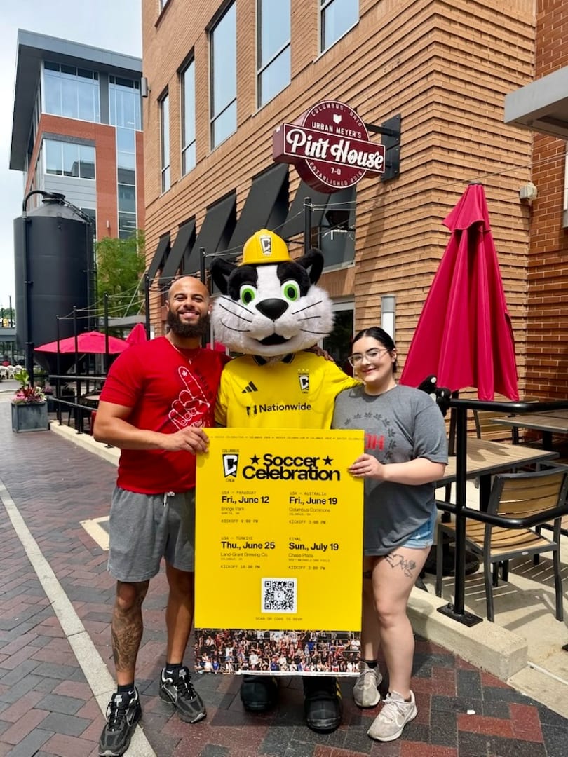 Two people and a cat mascot hold a "Soccer Celebration" sign outside Urban Meyer's Pint House.