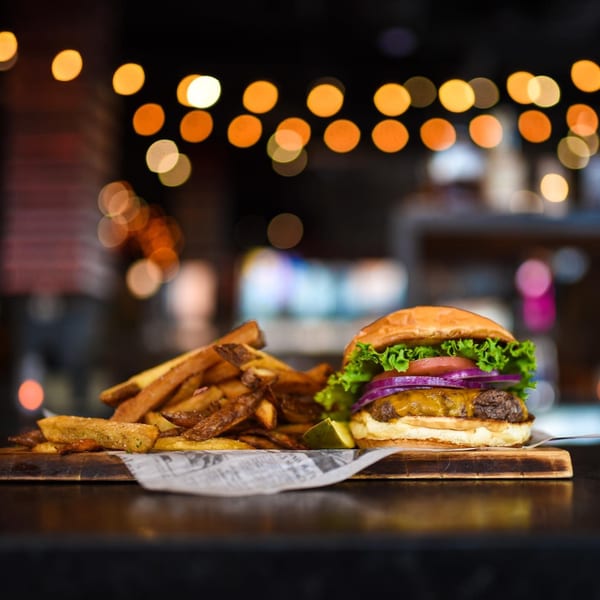 Cheeseburger with lettuce, tomato, red onion, and a side of fries and pickle on a wooden board.