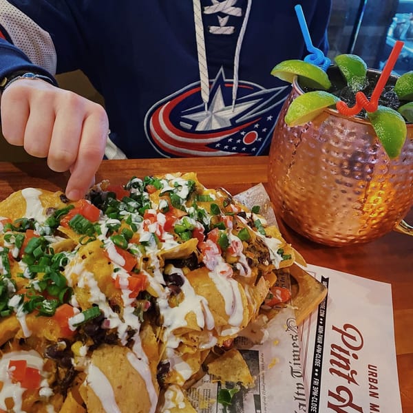 A person in a hockey jersey reaches for a large plate of nachos next to a copper mug cocktail.