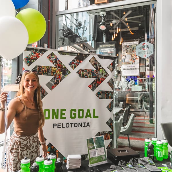 A smiling woman holds balloons next to a "One Goal Pelotonia" banner and merchandise table.