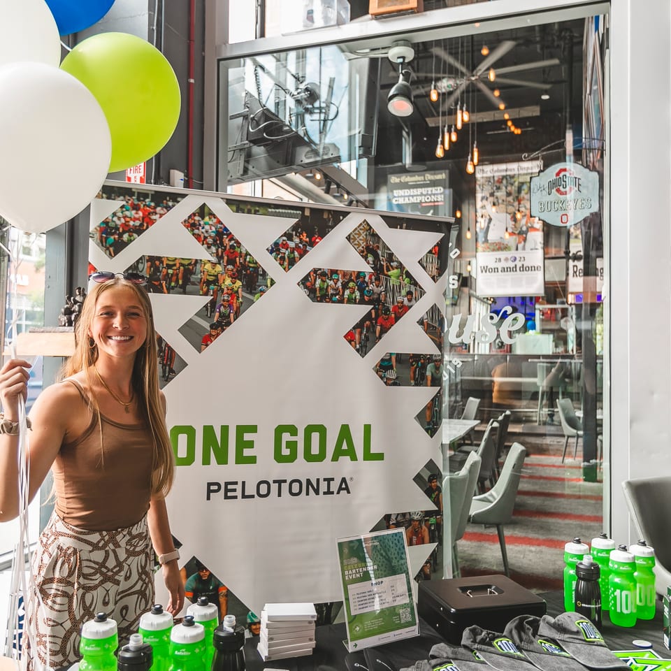 A smiling woman holds balloons next to a "One Goal Pelotonia" banner and merchandise table.