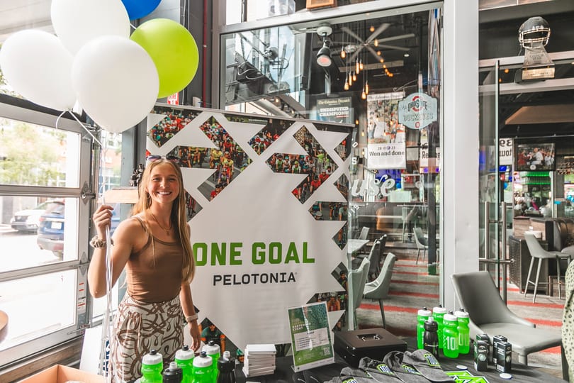 A smiling woman holds balloons next to a "One Goal Pelotonia" banner and merchandise table.