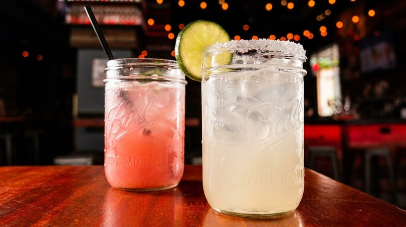 Two mason jar cocktails, one pink and one clear with a lime, on a wooden bar.