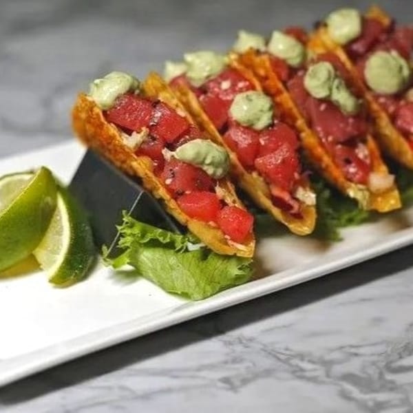 Four tuna poke tacos with avocado cream and lime wedges on a white rectangular plate.