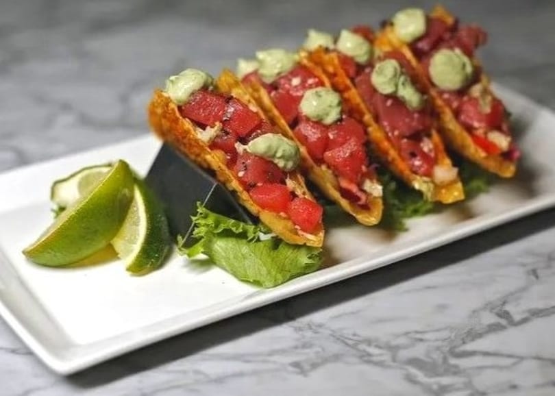 Four tuna poke tacos with avocado cream and lime wedges on a white rectangular plate.