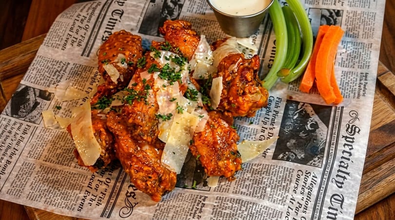 Spicy chicken wings with parmesan, parsley, celery, and carrots on newspaper-lined wooden board.