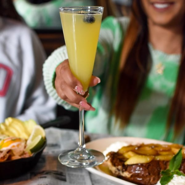 A person holds up a mimosa with blueberries, with food and another person blurred in the background.