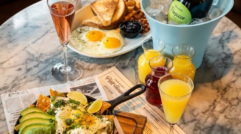 A marble table laden with brunch: huevos rancheros, fried eggs, toast, beans, and a mimosa bar.