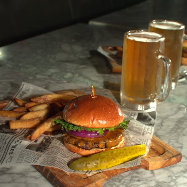 Cheeseburger with fries and a pickle spear on newspaper, with two mugs of beer on a marble table.