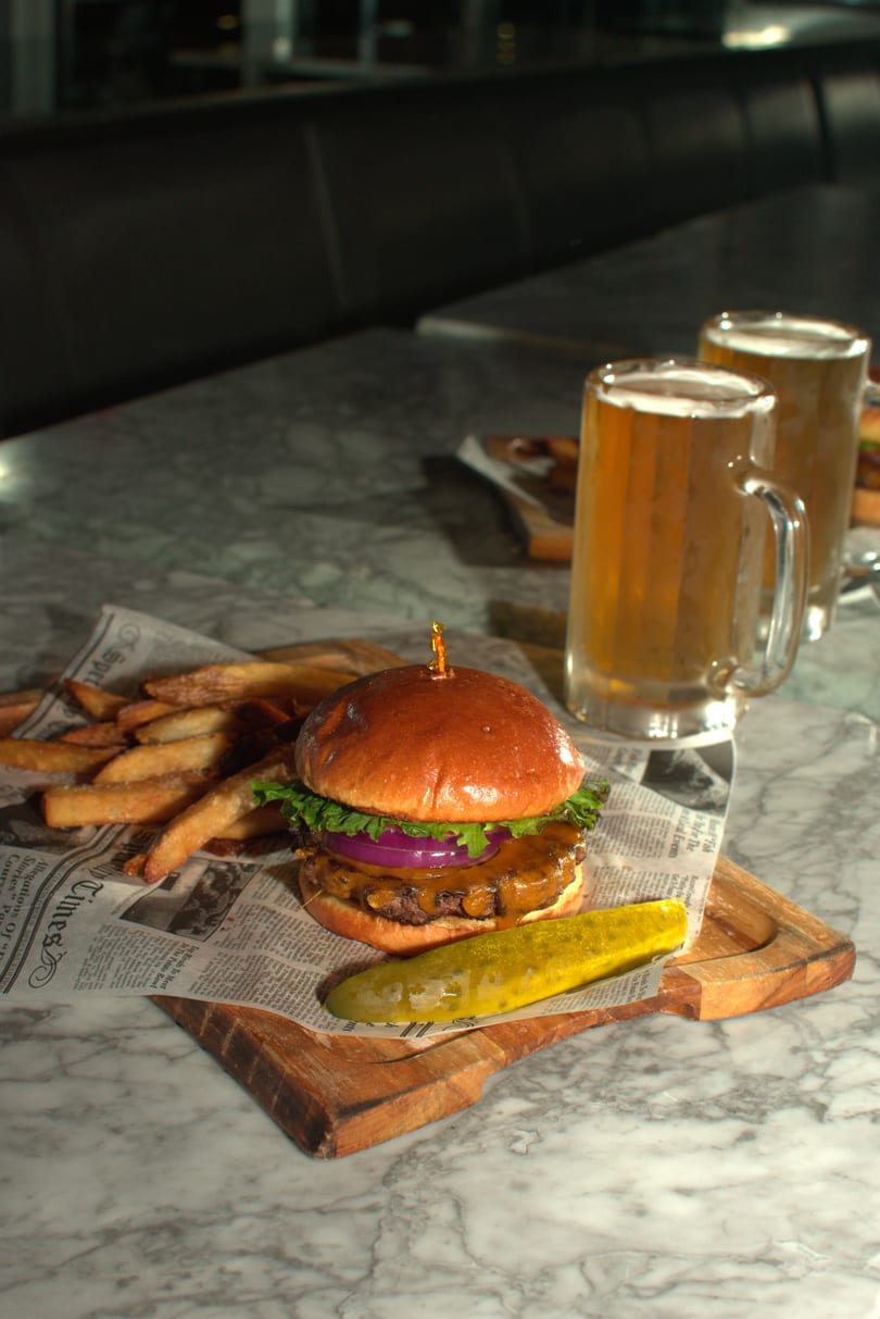 Cheeseburger with fries and a pickle spear on newspaper, with two mugs of beer on a marble table.