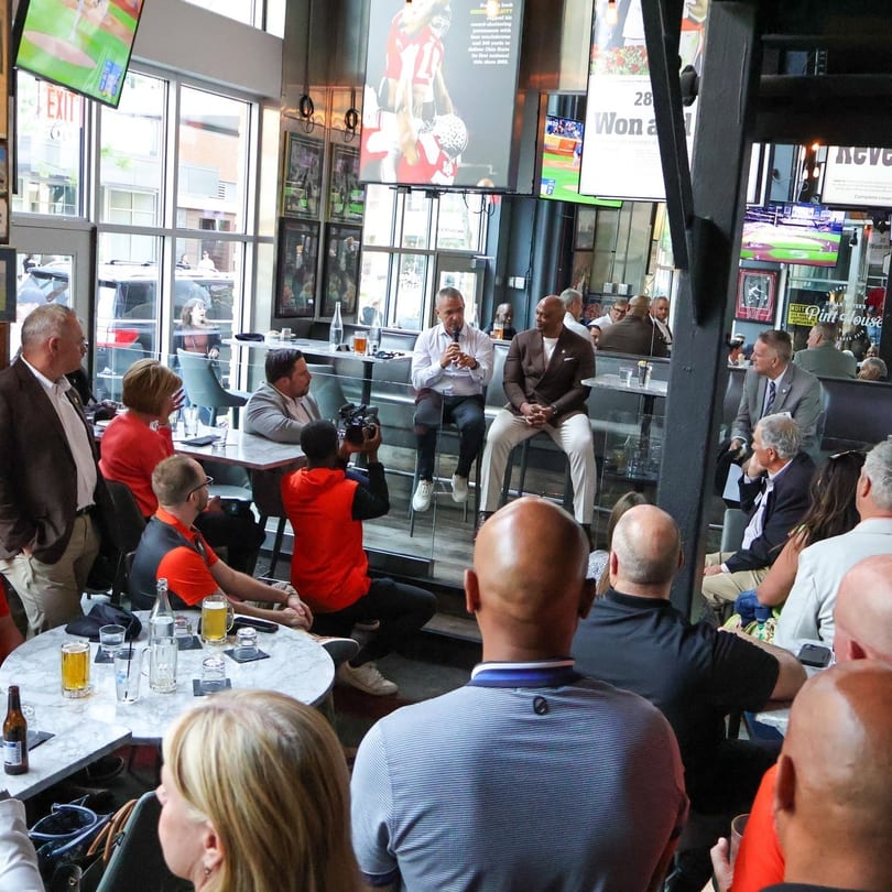 Two men speak on stools to a seated audience in a modern bar with multiple TVs.