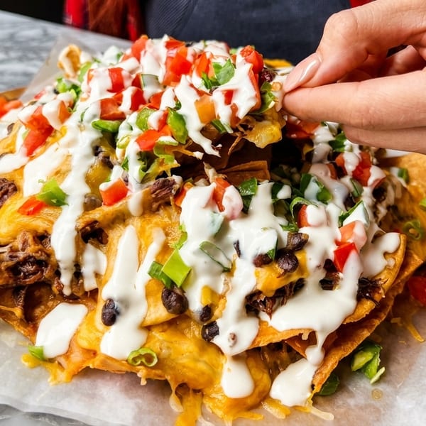 A hand reaches for a chip from a large platter of loaded nachos with cheese, meat, and toppings.