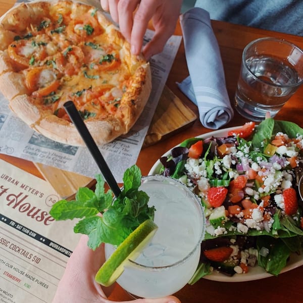 A hand reaches for a pizza, next to a salad, and a margarita with mint and lime.