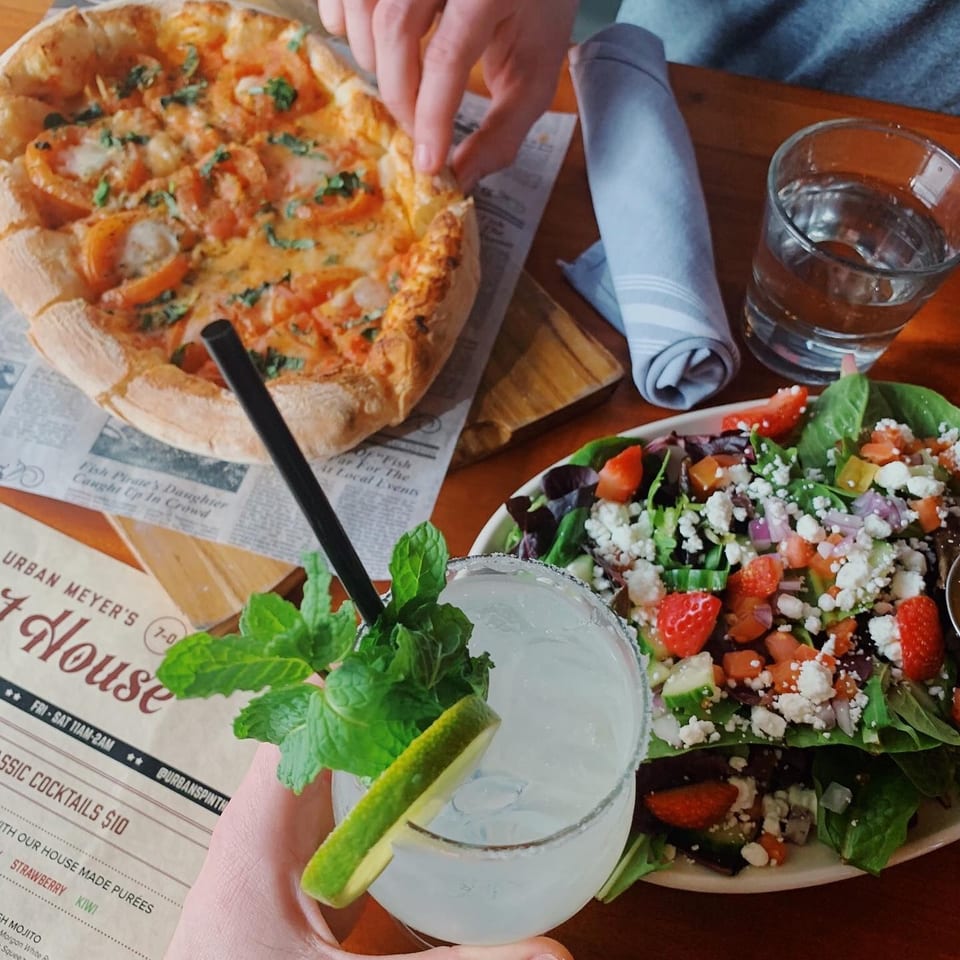 A hand reaches for a pizza, next to a salad, and a margarita with mint and lime.