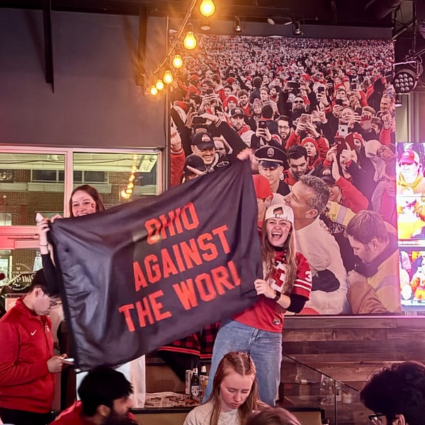 Two women holding a black "Ohio Against The World" flag in a lively restaurant with patrons.