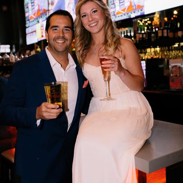 A smiling couple, a man holding a beer and a woman with champagne, at a bar.