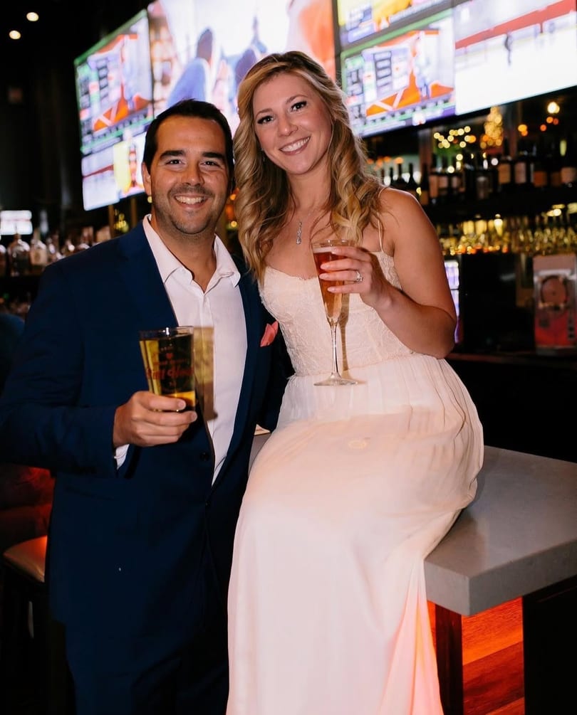 Smiling couple enjoying drinks at a lively bar with large screens in the background.